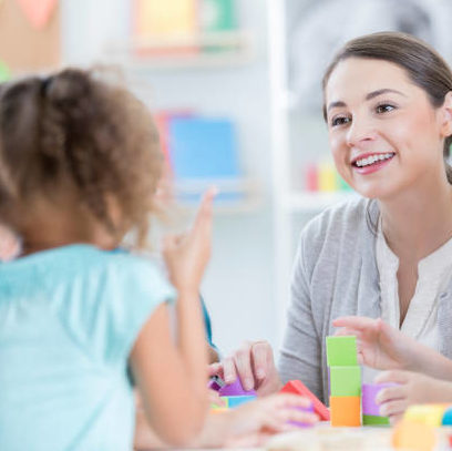 An attentive young kindergarten teacher sits at a table with her students in their classroom.  She looks up from playing with building blocks to listen to an unrecognizable student tell about her weekend.